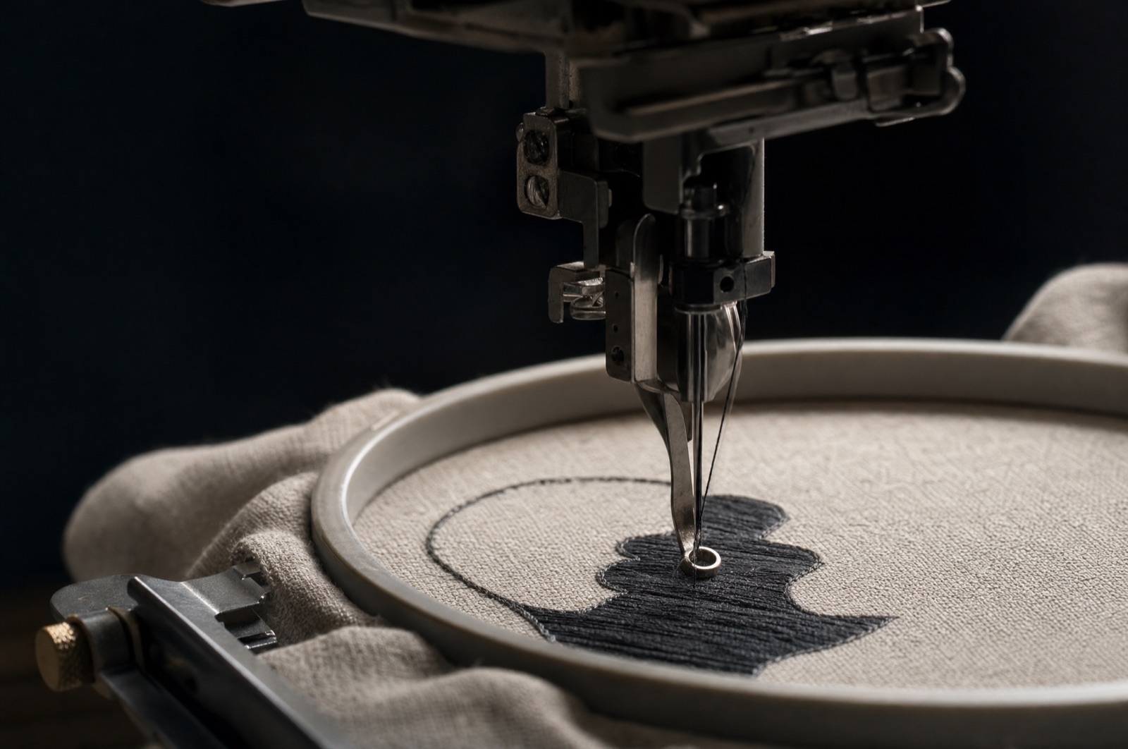 A macro view of a machine needle mid-stitch, finishing the satin edge of a dark appliqué shape in an embroidery hoop.
